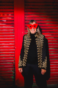 Bold black military-style jacket with gold trim, red masquerade mask, and red shutter backdrop at Harmonia NY