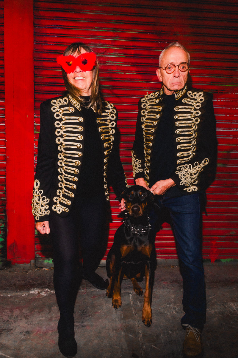 Bold festival wear from HARMONIA NY: couple in gold-embroidered jackets with dog, red backdrop